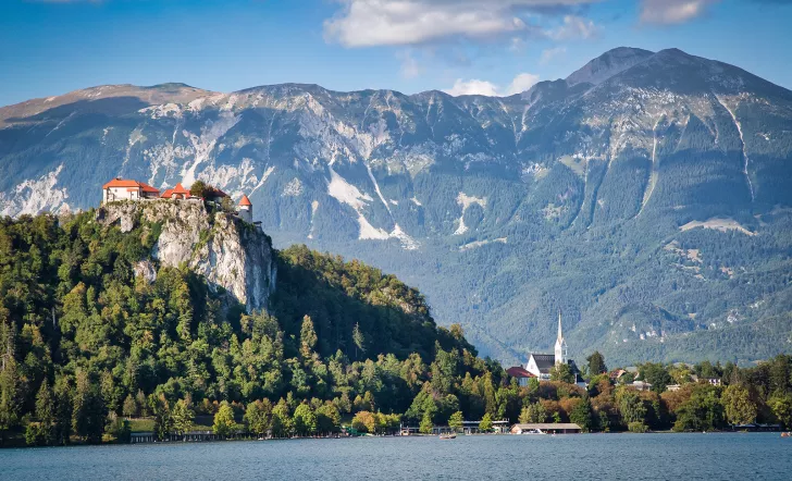 Wide shot of Bled Castle, town, lake below, mountain behind.