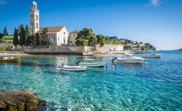 Wide shot of Hvar Island, white stone buildings, blue water, boats.