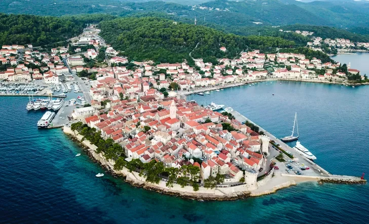 Wide shot of coastal Croatian town, white and tan buildings, boats, forest in distance. 