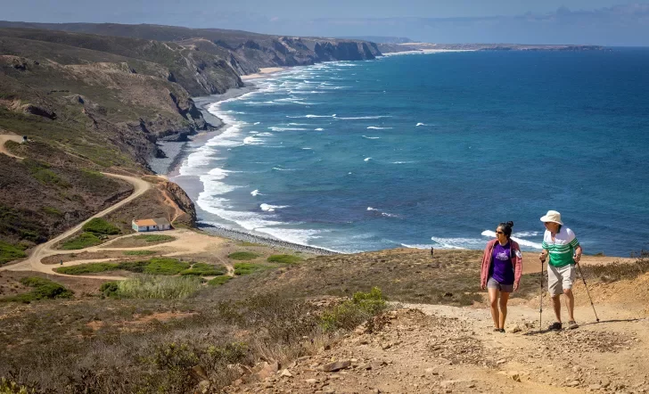 Two hikers walking up a sandy hill with the ocean in the background