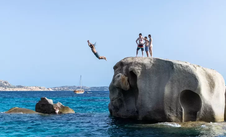 Four guests jumping off large rock, one in the water below.