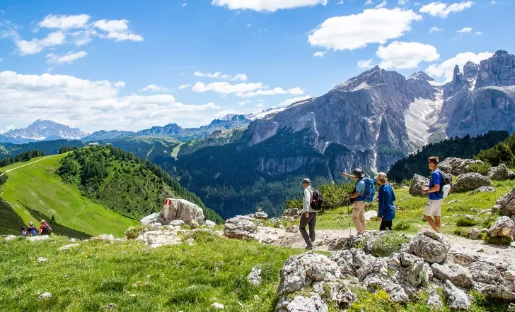 Group of guests hiking down mountain trail, Dolomite range in background.