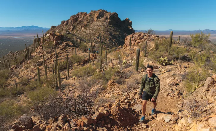 Man ascending a rocky, dirt trail with canyons and cacti behind him