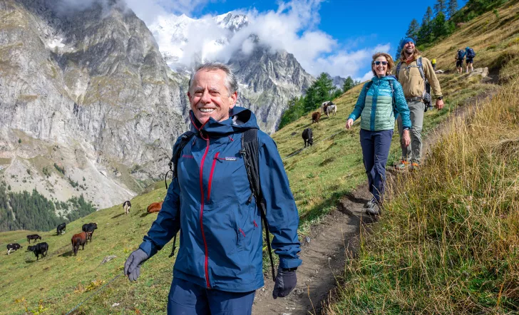Man hiking down a dirt path, with a large mountain and cows on a grass field in the background
