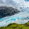 Shot of glacial vista, green cliff, blue ice, grey mountain.