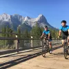 Two guests cycling on wooden path, Rockies in distance.