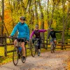 Three guests cycling down forest road, all smiling at camera, two signaling left turn.