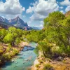 Wide shot of desert valley, blue stream, trees, cloudy sky.