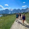 Group of guests walking next to small river, grassy meadow, mountains in background. 
