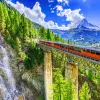 Wide shot of mountain train passing bridge, forest, mountains, sky in distance.