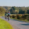Two people smiling while riding their bikes on a road surrounded by bushes and trees