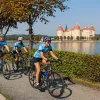 Three bikers on a road with a lake and large building in the background