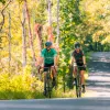 Man and woman riding bikes on an empty road, with tall trees surrounding the road