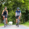 Man and woman smiling while riding their bikes on a road, with large trees surrounding