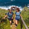 Two women walking up stairs on a grassy hill, with the ocean in the background