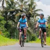 Two women smiling while ridings bikes on a road surrounded by tall plants
