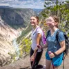 Two girls using hiking poles, walking on a dirt trail and large mountains in the distance