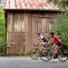 Man and woman wearing biking gear, riding bikes in front of a wooden barn on a road