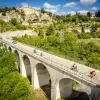 An aerial view of several cyclists crossing a large stone arch bridge above a wooded valley. A historic hilltop village with stone buildings sits on rocky cliffs in the background under a blue sky with scattered clouds.