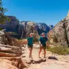 Two hikers walk along a rocky trail in Zion National Park, surrounded by towering sandstone cliffs, canyon walls, and desert vegetation beneath a clear blue sky.