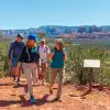 Group of hikers walking along a red dirt trail overlooking Sedona’s red rock formations and desert landscape under a clear blue sky.