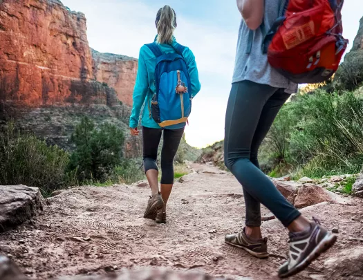 Two guests walking down desert trail, orange cliffs, bushes beside them.