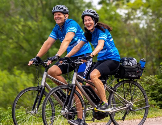 Two guests biking on forest road, smiling at camera.