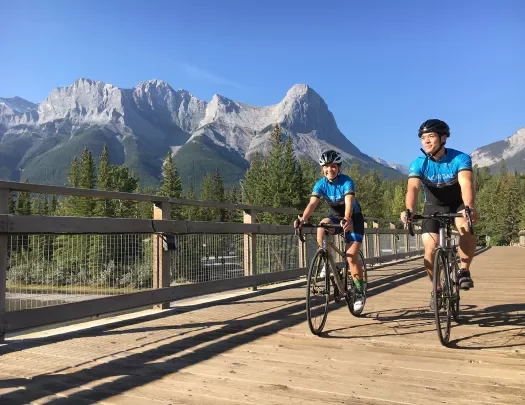 Two guests cycling on wooden path, Rockies in distance.