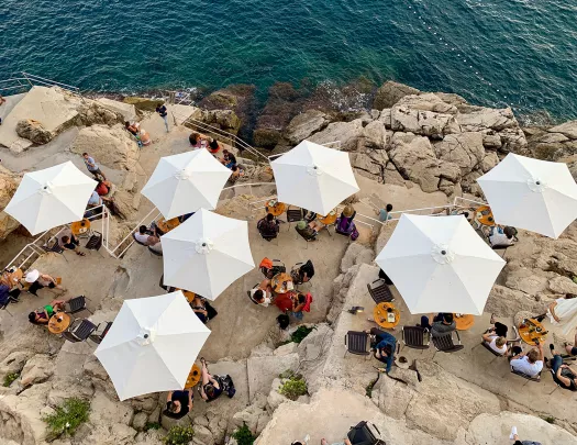 Overhead shot of people eating beneath white umbrellas, stone cliffs, ocean.