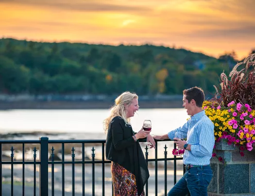 Two guests chatting, wine glasses in hand, sunset and beach in background.