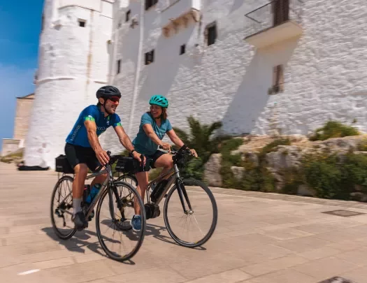Motion shot of two guests cycling next to large white brick wall.