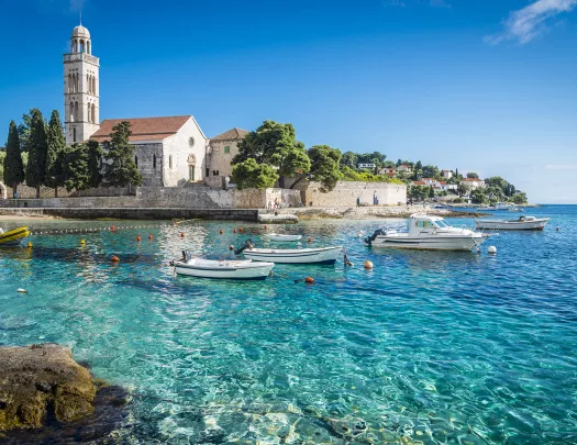 Wide shot of Hvar Island, white stone buildings, blue water, boats.