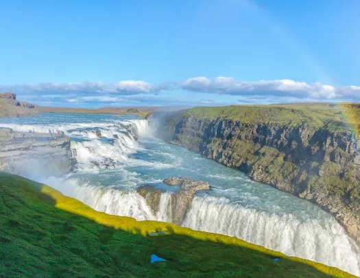 Wide shot of alpine vista, waterfall, rainbow, cliffs on either side. 