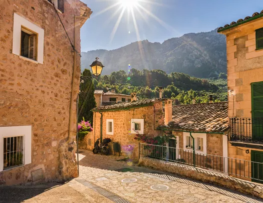 POV shot in tan stone village, sun shining, forested mountains in distance.