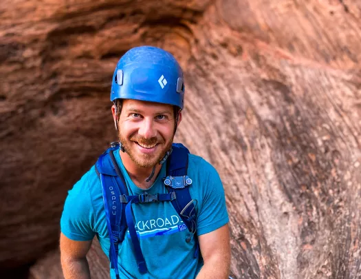 Guest in rock climbing gear on cliffside, smiling at camera.