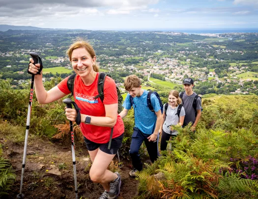 Four guests walking up hillside trail, French village, coast in distance. 