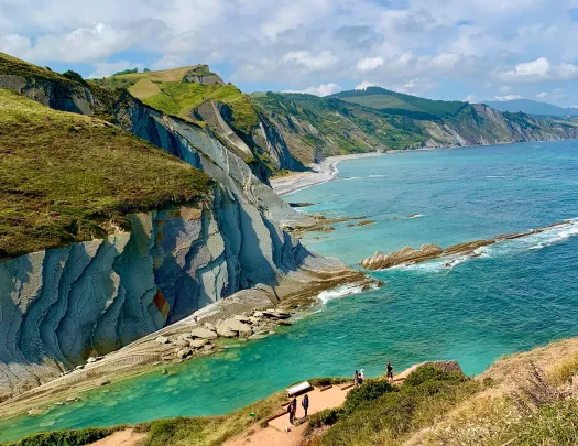 Wide shot of rocky coastline. Blue water, layered rocks, guests in distance.