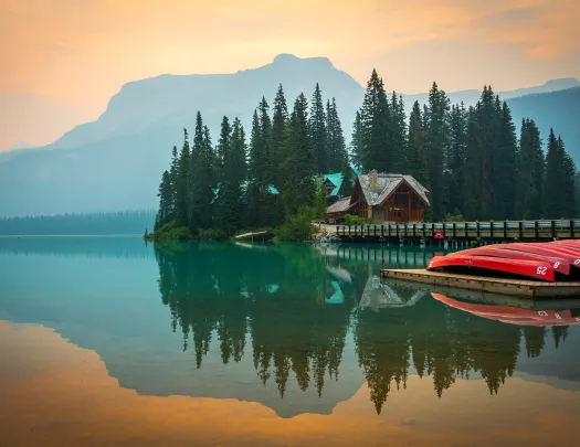 Wide shot of large lake, wooden house, trees, canoes, mountains visible.