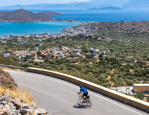 Guest cycling on road, arid coastal town in distance, ocean behind it.