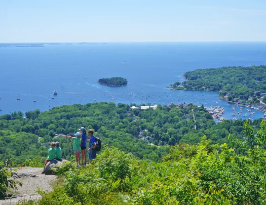 Wide shot of a coastal vista, sailboats visible in water, group of young guests in foreground.