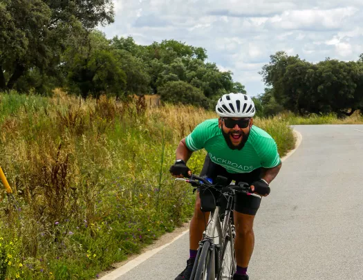 Guest on road cycling towards camera, smiling, trees, clouds behind.