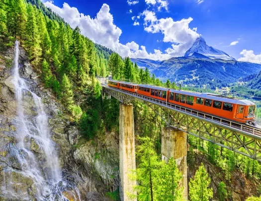 Wide shot of mountain train passing bridge, forest, mountains, sky in distance.