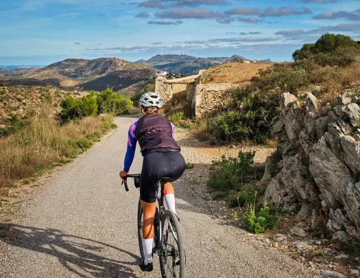 Guest cycling down gravel road, towards stone ruins, golden hills. 