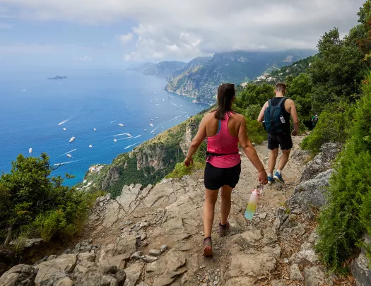Two guests hiking down cliffside, boats in ocean down below.