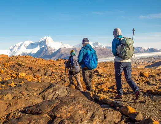 Three guests walking over orange rocks, snowy peaks in distance.