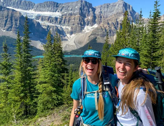 Two guests smiling for camera, snowy mountain, trees, blue sky in background.