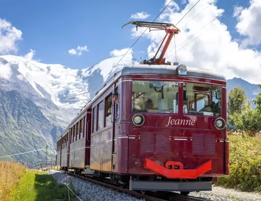 Shot of a red train, "Jeanne", mountains & clouds behind it.
