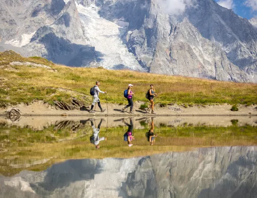 Three guests walking past reflective lake, meadow, snowy mountain in distance. 