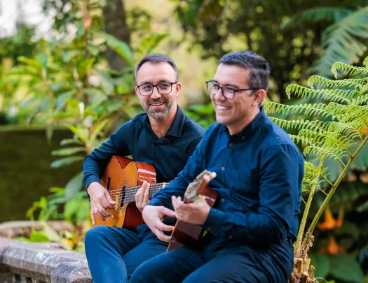 Two men sitting on a stone fence playing guitars and smiling