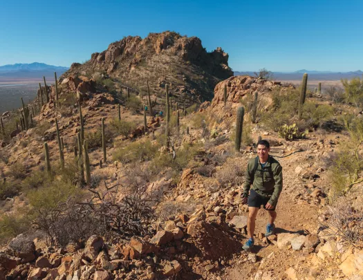 Man ascending a rocky, dirt trail with canyons and cacti behind him